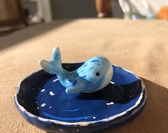 May include: A small, handcrafted ceramic whale figurine in shades of blue, resting on a dark blue dish with white speckles. The dish has a raised edge, creating a miniature ocean scene. The whale is approximately 2 inches long.