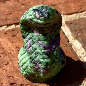 May include: Green and purple serpentine stone carved into a cobra statue. The statue is sitting on a brick surface.
