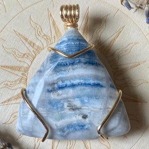 May include: A triangular pendant made of blue and white banded agate, wrapped in gold wire. The pendant is sitting on a wooden surface with a circular design.