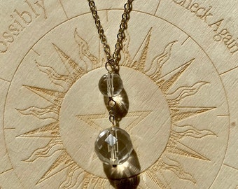May include: A gold chain necklace with two clear crystal beads hanging from it. The necklace is lying on a wooden surface with a circular design.