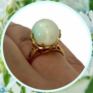May include: A gold-colored ring featuring a large, pale green sphere. The ring's band has an ornate, openwork design that supports the sphere. The sphere appears to be a gemstone or a decorative element.
