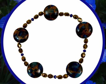 Puede incluir: Una pulsera de cuentas con cinco piedras grandes, redondas, de color azul oscuro y marrón. La pulsera está ensartada con cuentas doradas y azules más pequeñas. La pulsera está sobre un fondo blanco con un borde azul y flores azules.