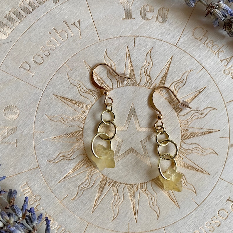 May include: A pair of gold earrings with three small gold hoops and a light yellow crystal charm on each earring.
