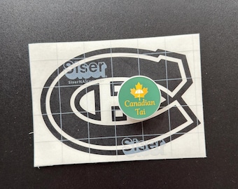Op de afbeelding: Een ronde groene sticker met een geel esdoornblad, een klein wit paraplu-icoontje en de tekst Canadian Tai in een geel lettertype, geplaatst op een rechthoekig wit transfervel met een zwarte afbeelding van een grote letter C.