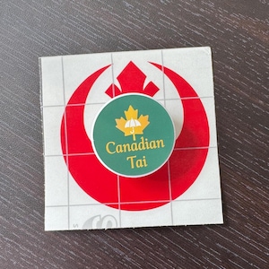 Peut inclure: Un autocollant circulaire sur fond vert avec une feuille d'érable jaune contenant une icône de parapluie blanc et le texte Canadian Tai en jaune, superposé sur un support carré blanc avec un grand symbole rouge de l'Alliance Rebelle.