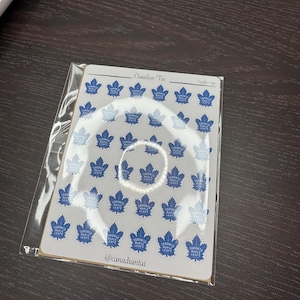 Könnte beinhalten: Ein Bogen mit kleinen, blauen Toronto Maple Leafs Logo-Aufklebern, die in einem Raster auf einem weißen Träger angeordnet sind. Oben auf dem Bogen steht der Text Canadian Tai und Leafs-xs, unten der Social-Media-Name @canadiantai.