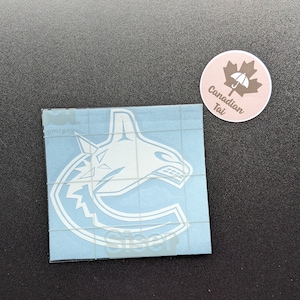 May include: A white vinyl decal featuring the Vancouver Canucks orca logo on a light blue transfer sheet with a grid pattern. Next to it is a small round sticker with a brown maple leaf, a white umbrella icon, and the text Canadian Tai.