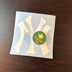 May include: A white vinyl decal featuring the interlocking NY logo on a grid-lined transfer sheet, with a small circular green sticker placed on top. The green sticker displays a yellow maple leaf icon and the text Canadian Tai in yellow font.