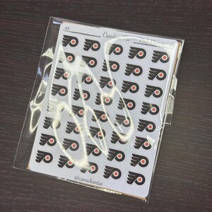 May include: A clear plastic sleeve containing a sheet of small, circular stickers. Each sticker features the black and orange Philadelphia Flyers logo. The top of the sheet is labelled 63, Canadian Tai, and Phil Flyers - xs, with @canadiantai at the bottom.