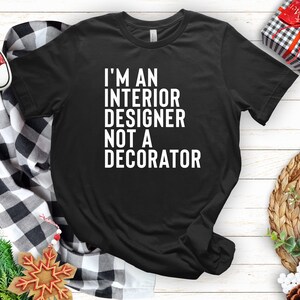 May include: Black t-shirt with the text "I'M AN INTERIOR DESIGNER NOT A DECORATOR" in white. The shirt is on a white wooden surface, with a black and white plaid scarf, red sneakers, and holiday decorations.