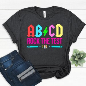May include: A dark grey t-shirt with a colorful graphic that says "ABC Rock the Test". The graphic features a lightning bolt and colorful pencils.