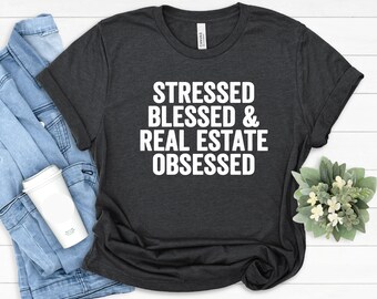 Könnte beinhalten: Dunkelgraues T-Shirt mit dem weißen Text "STRESSED BLESSED & REAL ESTATE OBSESSED". Eine Jeansjacke und ein weißer Reisebecher sind im Hintergrund zu sehen. Eine kleine Pflanze ist ebenfalls sichtbar. Das T-Shirt ist gefaltet.