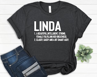 Può includere: Una maglietta grigio scuro con la parola "LINDA" stampata in bianco. Il testo sotto definisce il nome come "Una donna bella, intelligente, forte, che realizza la sua grandezza. Elegante, sfrontata e un po' furba."