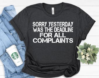 May include: Dark gray t-shirt with the text "SORRY YESTERDAY WAS THE DEADLINE FOR ALL COMPLAINTS" in white block letters. The shirt is folded and placed on a light blue denim jacket. A Starbucks coffee cup and a small floral arrangement are also in the image.