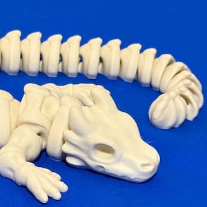 May include: A cream-colored articulated lizard skeleton toy. The toy features a detailed design of a lizard skeleton with a segmented body, legs, and a head with eye sockets. The toy is posed on a blue surface.