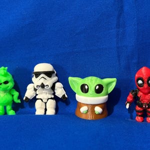 May include: Four small figurines of popular characters. The Grinch is green, a Stormtrooper is white and black, Baby Yoda is green and brown, and Deadpool is red and black. They are arranged on a blue surface.