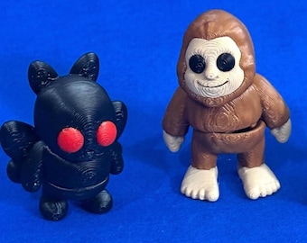 May include: Two 3D-printed figurines on a blue background. One is a black creature with red eyes and bat-like wings. The other is a brown Bigfoot-like figure with a light beige face and feet. Both are standing upright.