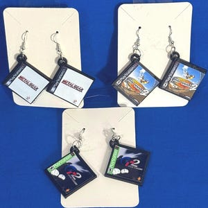 May include: Earrings featuring miniature PlayStation game cases. The earrings showcase game titles like "Metal Gear", "Tony Hawk's Pro Skater 2", and "Gran Turismo 2". Each earring has a silver hook and a black square pendant.
