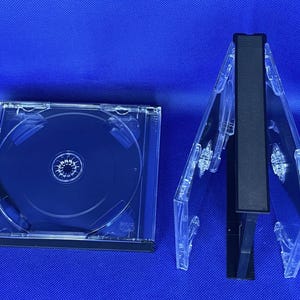 Könnte beinhalten: Zwei transparente CD-Hüllen aus Kunststoff, eine ist geschlossen und die andere ist geöffnet mit einer schwarzen CD darin. Die geöffnete Hülle ist in der Mitte gefaltet.