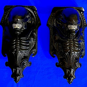 Puede incluir: Dos apliques de pared negros con un diseño detallado de Alien. Cada aplique presenta una cabeza y un cuerpo de Alien esculpidos, con intrincadas nervaduras y un marco decorativo. Los apliques son de un material oscuro, posiblemente metálico, sobre un fondo azul.