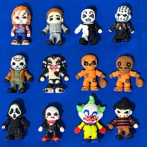 May include: A collection of twelve miniature horror movie character figurines. The figures include Chucky, Michael Myers, a mime, Jason Voorhees, the Predator, Sam, Ghostface, Billy the Puppet, a clown, and Freddy Krueger. Each figure is brightly colored and detailed.