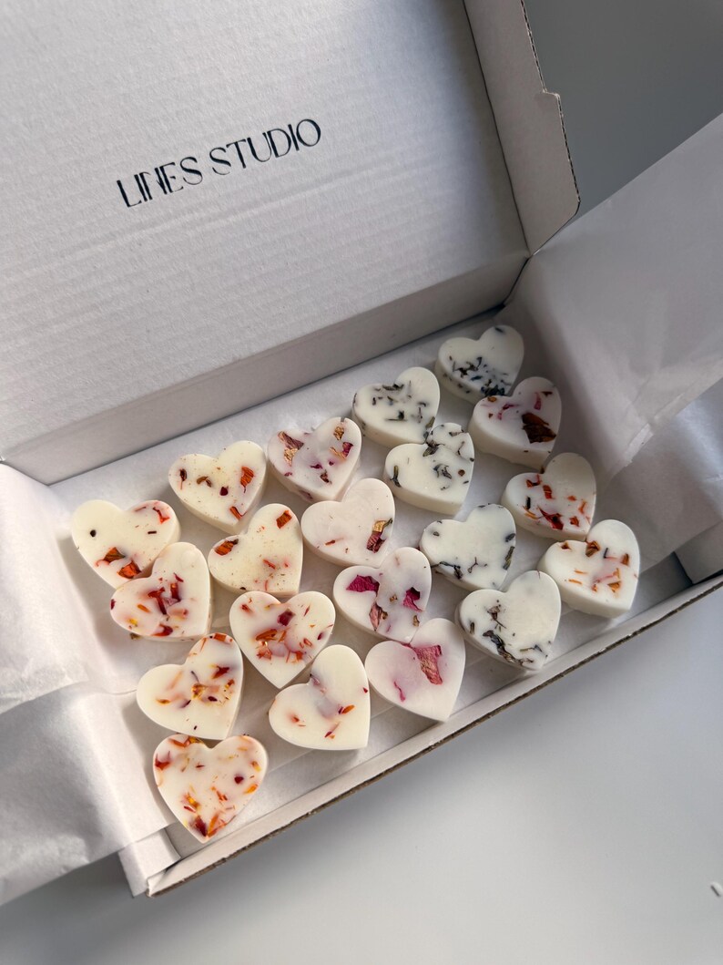 May include: A white box with the text "LINES STUDIO" contains heart-shaped wax melts. The melts are white, decorated with dried flower petals in shades of red, pink, and black. The box is open, revealing the contents.