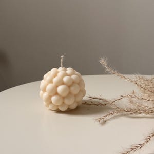 Könnte beinhalten: Eine kugelförmige, cremefarbene Kerze mit einer strukturierten, blasenartigen Oberfläche. Die Kerze hat einen einzelnen Docht und steht auf einer hellen Oberfläche. Getrocknetes Ziergras befindet sich im Hintergrund.