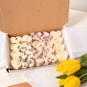 May include: A cardboard box filled with heart-shaped wax melts, each decorated with dried flowers and botanicals. The melts are arranged on white paper. Yellow tulips are in the foreground.