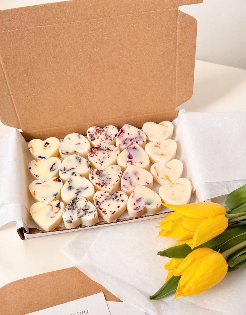 May include: A cardboard box filled with heart-shaped wax melts, each decorated with dried flowers and botanicals. The melts are arranged on white paper. Yellow tulips are in the foreground.