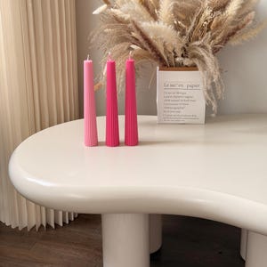 Peut inclure: Trois bougies effilées, hautes et fines, présentant des nuances de rose allant du pastel clair au magenta profond, chacune surmontée d'une mèche blanche. Les bougies sont disposées en rangée et présentent une texture cannelée verticale.