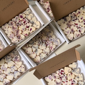 May include: Several boxes filled with heart-shaped, off-white wax melts, some adorned with red and purple floral accents. The boxes are brown with the text "LINES STUDIO" printed on them. The melts are arranged in white trays.