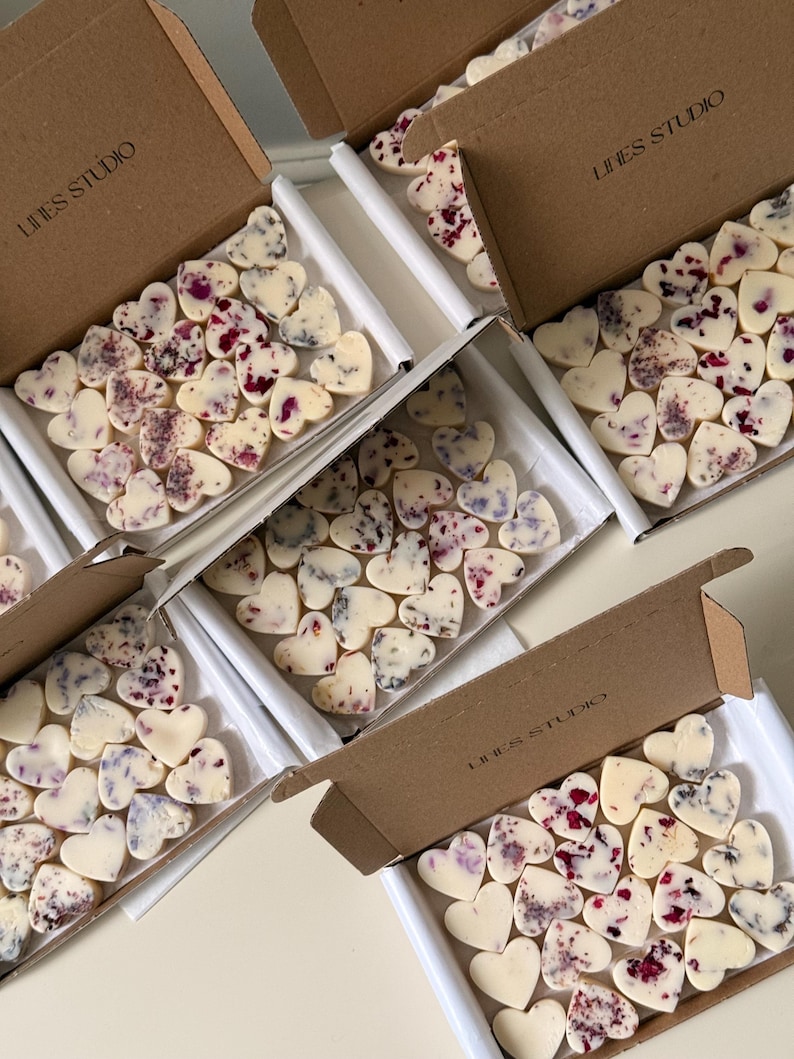May include: Several boxes filled with heart-shaped, off-white wax melts, some adorned with red and purple floral accents. The boxes are brown with the text "LINES STUDIO" printed on them. The melts are arranged in white trays.