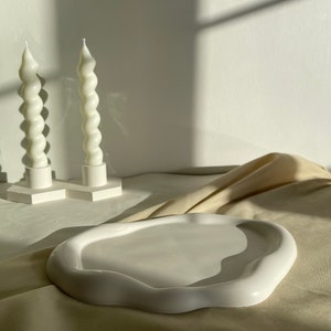 Puede incluir: Una bandeja de cerámica blanca con bordes ondulados y superficie lisa. Dos velas blancas en espiral sobre bases cuadradas en el fondo. Los objetos están dispuestos sobre una tela de tono neutro.