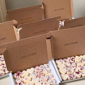 May include: Heart-shaped wax melts with floral accents are displayed in open cardboard boxes. The boxes are labeled "LINES STUDIO". The melts are white, with some decorated with red and purple petals.