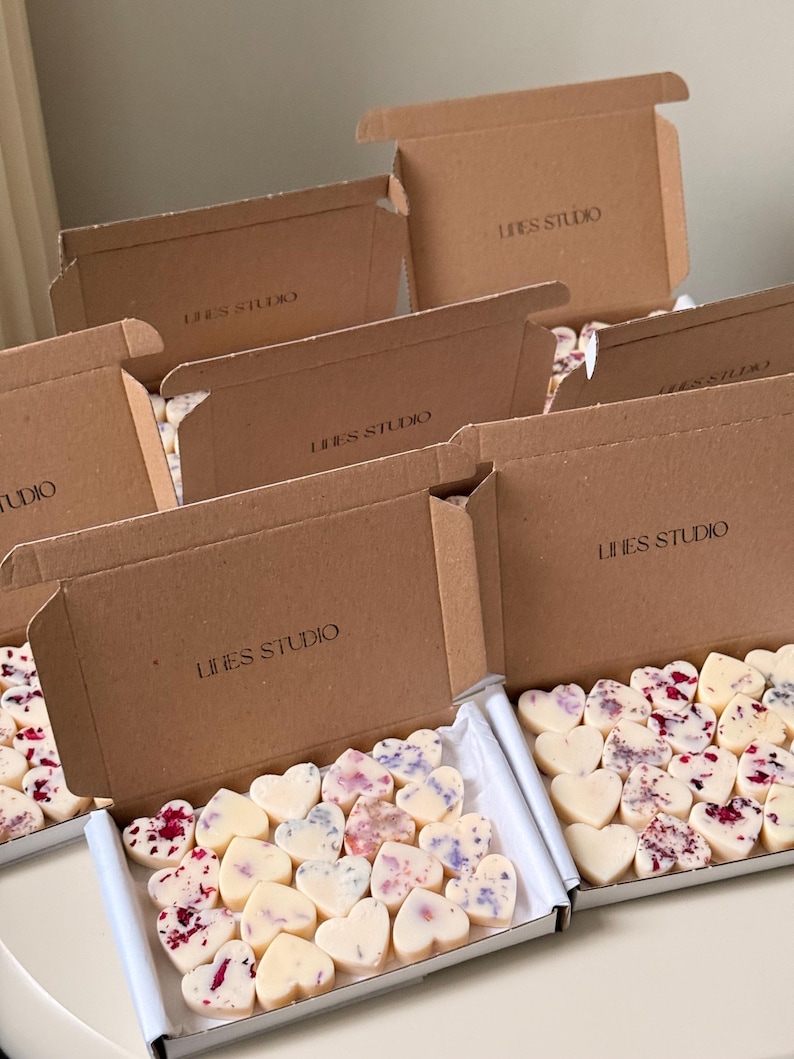 May include: Heart-shaped wax melts with floral accents are displayed in open cardboard boxes. The boxes are labeled "LINES STUDIO". The melts are white, with some decorated with red and purple petals.