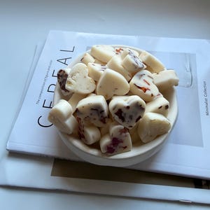 May include: A white ceramic bowl filled with heart-shaped wax melts. Some melts have red and brown inclusions. The bowl sits on a magazine with the word "CEREAL" and the text "The composition of figure and color" visible.