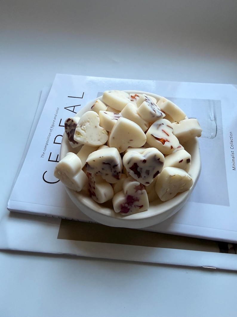 May include: A white ceramic bowl filled with heart-shaped wax melts. Some melts have red and brown inclusions. The bowl sits on a magazine with the word "CEREAL" and the text "The composition of figure and color" visible.