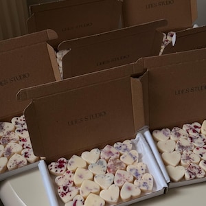 May include: Several open cardboard boxes filled with heart-shaped wax melts. The melts are white and decorated with dried flowers. The boxes are labeled "LINES STUDIO".