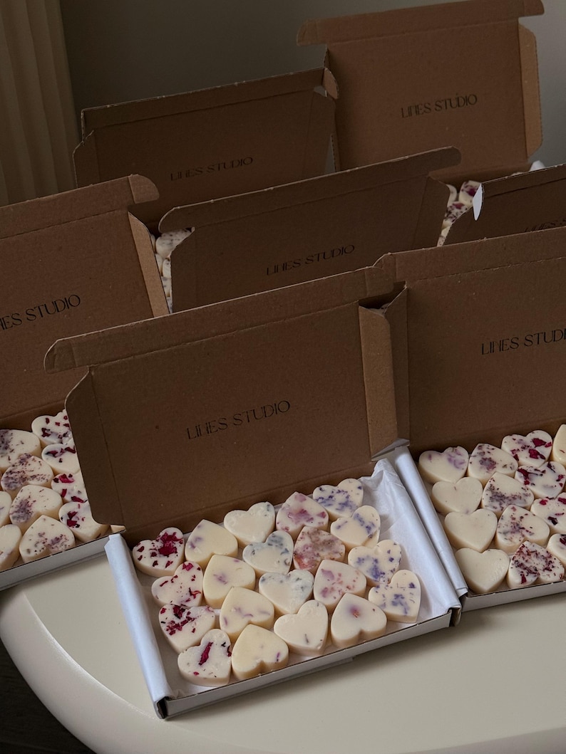 May include: Several open cardboard boxes filled with heart-shaped wax melts. The melts are white and decorated with dried flowers. The boxes are labeled "LINES STUDIO".