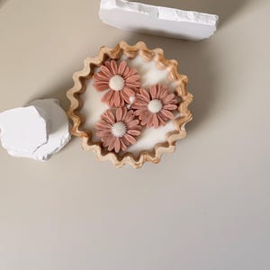 May include: A decorative candle featuring a white wax base and three pink daisy-shaped accents. The candle sits in a round, scalloped-edge holder. Two white, irregularly shaped stones are placed nearby on a neutral surface.