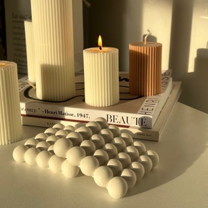 Può includere: Una collezione di candele decorative in varie dimensioni e colori. Le candele sono cilindriche con dettagli a coste verticali. Una candela è accesa, mostrando una fiamma calda. Un oggetto decorativo bianco e sferico è in primo piano. I libri sono sullo sfondo.