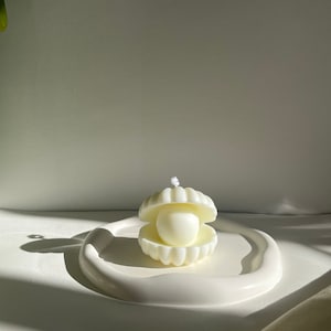 May include: A white clam shell candle with a pearl inside, resting on a white, curved tray. The candle has a wick and is unscented. The design is minimalist and elegant, suitable for home decor.