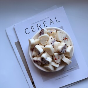 May include: A white ceramic bowl filled with heart-shaped, white-coated candies with red and brown inclusions. The bowl rests on a magazine with the word "CEREAL" in large letters. The magazine has the text "The composition of figure and color".