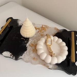 May include: A decorative tray with gold handles, featuring a black and white marbled design. It holds a white candle, a gold watch, and a white, bubble-shaped dish. Gold flakes add a touch of elegance.
