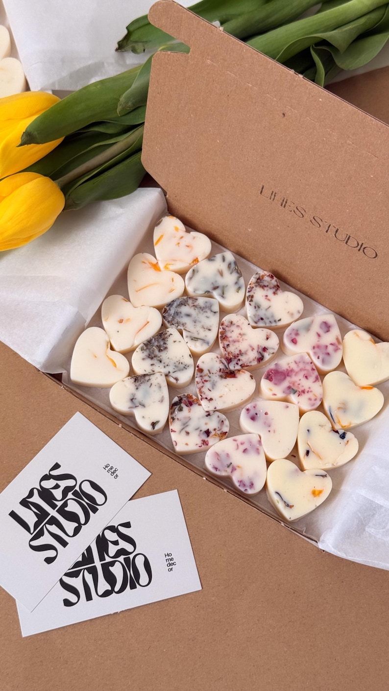 May include: A box of heart-shaped wax melts with floral inclusions. The melts are white and various colors, including orange, purple, and gray. The box is brown, and the brand name "LINES STUDIO" is printed on the box and on two white cards.