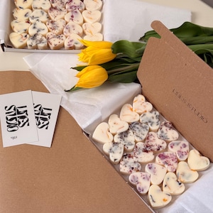 May include: Two cardboard boxes filled with heart-shaped wax melts, some with floral inclusions. A bouquet of yellow tulips and two business cards are also present. The boxes are on a light-colored surface.