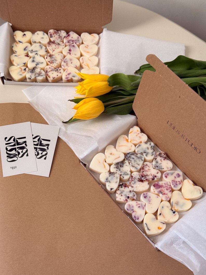 May include: Two cardboard boxes filled with heart-shaped wax melts, some with floral inclusions. A bouquet of yellow tulips and two business cards are also present. The boxes are on a light-colored surface.