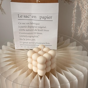 May include: A cream-coloured bubble candle with a white wick rests on a decorative paper fan. Behind it is a white paper bag with French text, including "Le sac en papier". Dried plants are in the background.