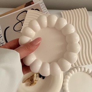Op de afbeelding: Een verzameling witte decoratieve trays met unieke ontwerpen. Eén tray heeft een bubbelrand, een andere een kralenrand en een derde een golvend patroon. Een goudkleurig horloge en een zwarte strik zijn zichtbaar op een van de trays.
