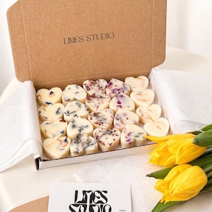 May include: A box of heart-shaped wax melts with floral inclusions. The melts are arranged in a white box, and the box is inside a larger cardboard box. The text "LIMES STUDIO" is printed on the cardboard box and on a business card. Yellow tulips are in the foreground.